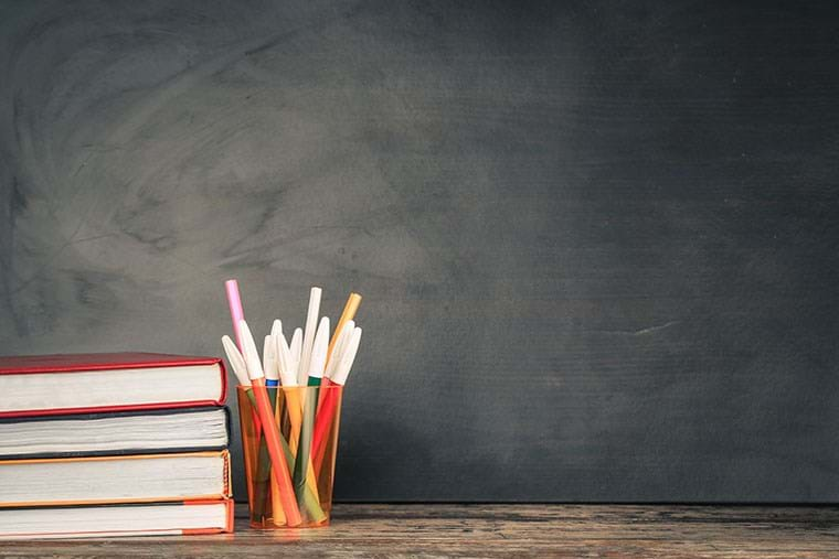 A stack of pens and books sits before a chalkboard