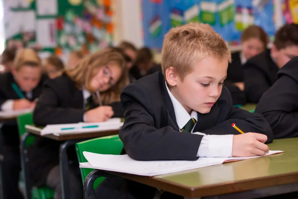 A young boy in a school uniform writing at his desk in a classroom.