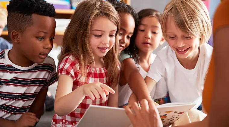 A group of young children look at a picture book with their teacher.