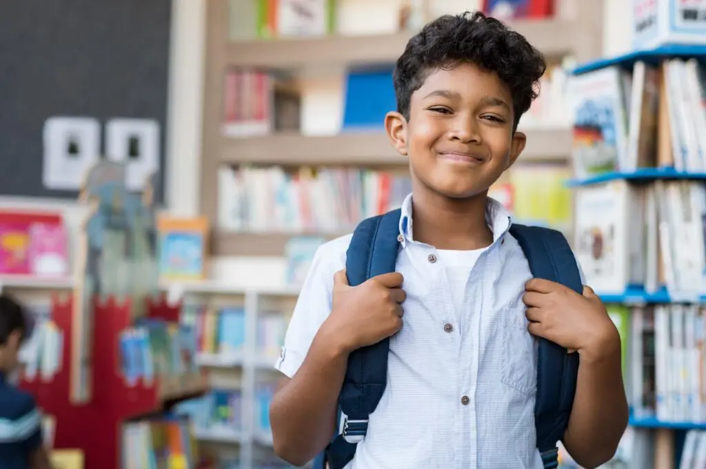 Smiling schoolboy with backpack in front of bookshelves.