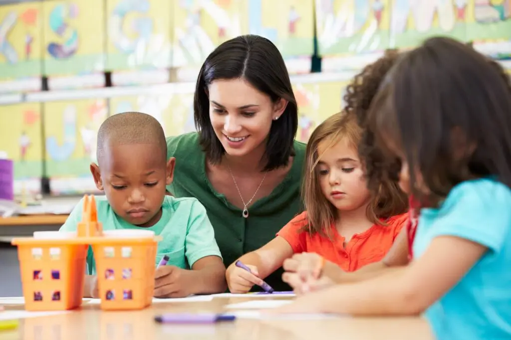 Teacher assisting young children with coloring in a classroom.