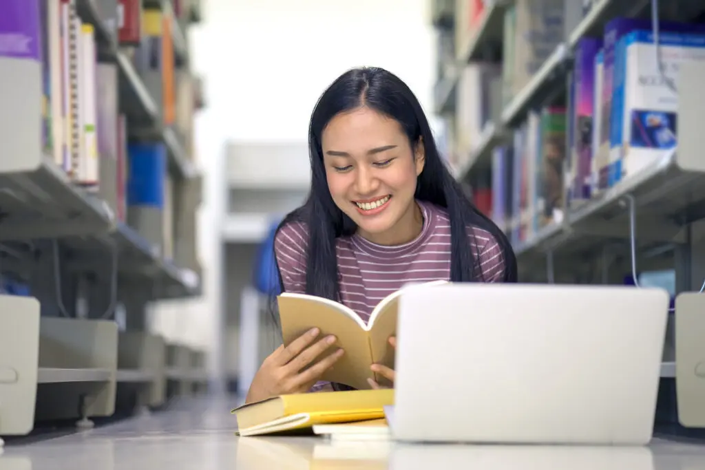 Smiling student reading a book on the library floor with a laptop nearby.